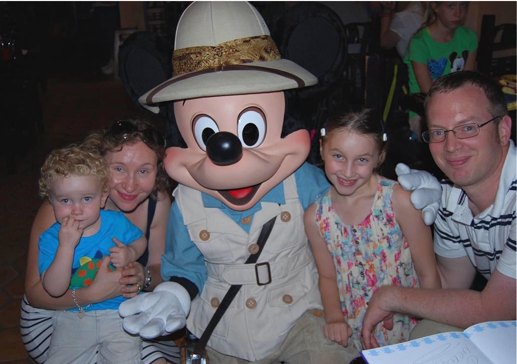 Crouching woman with toddler in her arms, a young girl and a man next to her and Mickey Mouse in the middle, all very happy 